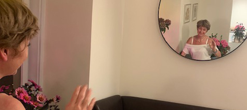 Raffaella Correngia, Geschäftsführerin, lächelt in einem runden Spiegel und winkt. Sie befindet sich in einem gemütlich dekorierten Raum mit Blumen im Hintergrund.