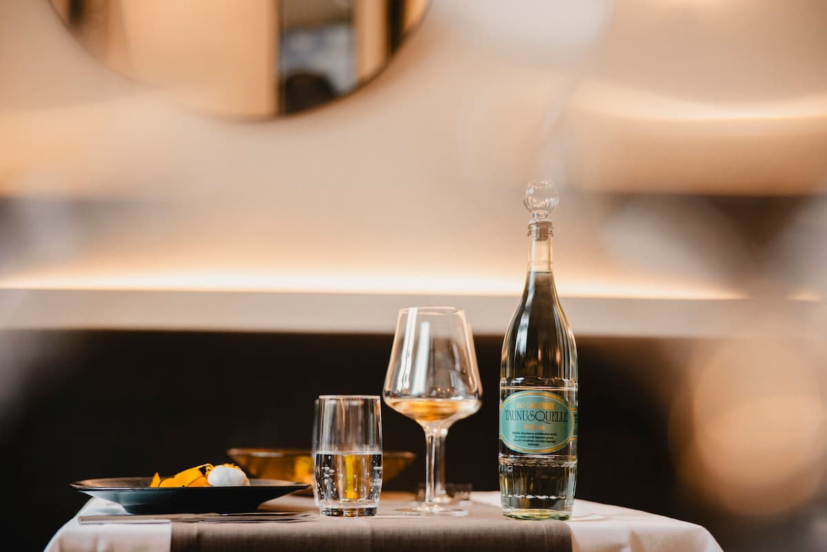 Elegantes Tischgedeck: Tauni&Quell Wasserflasche mit Kristallgläsern und Gourmet-Dessert in warmem, sanft beleuchtetem Ambiente